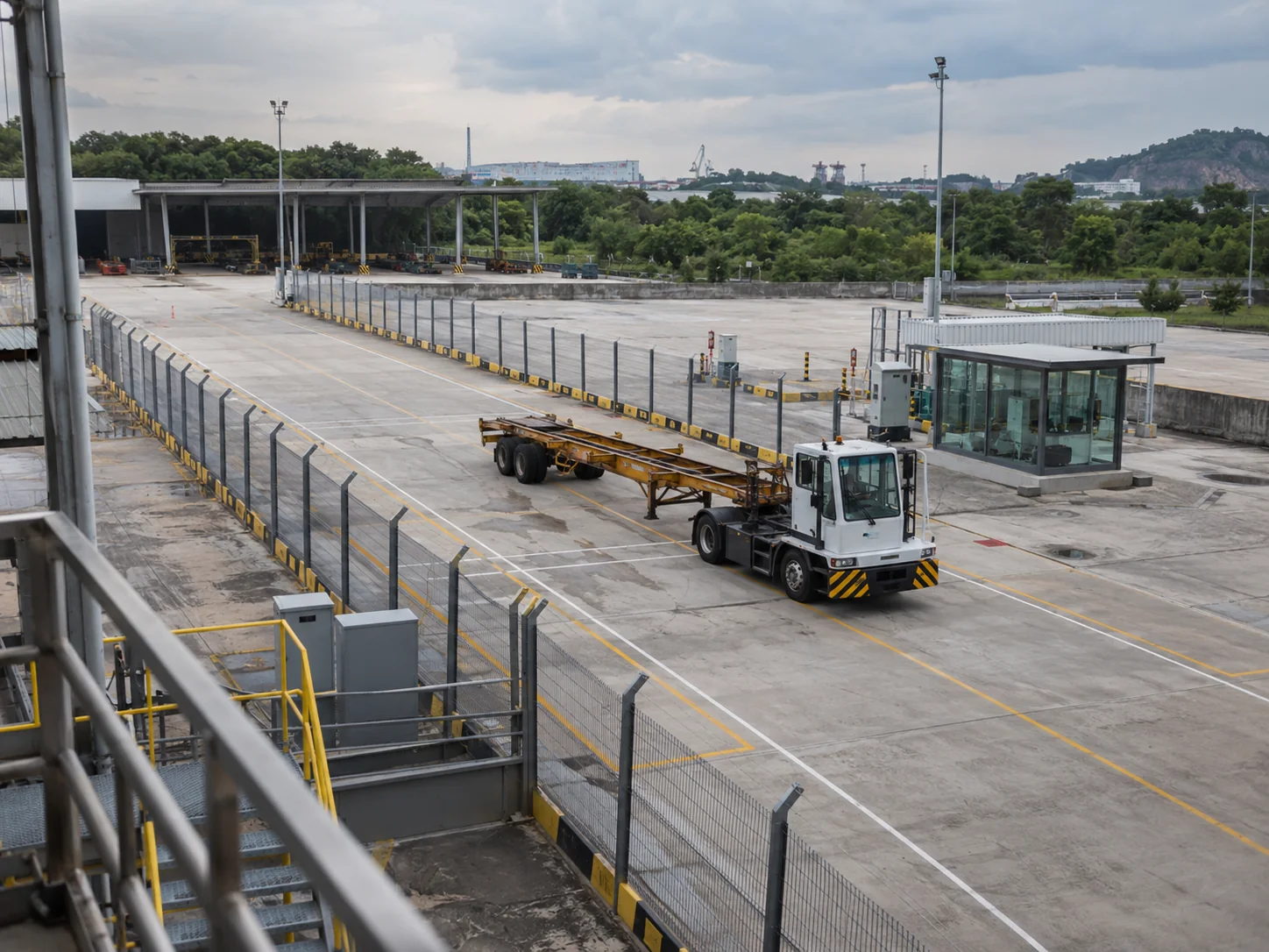 AI-generated illustration. High-angle wide editorial photograph from a warehouse mezzanine overlooking a quiet generic industrial test yard in Malaysia, one ordinary unbranded terminal tractor with a short empty trailer parked on a clearly marked fenced test lane, a small glazed control hut visible well away from the vehicle. Overcast natural light, functional worn concrete, no driverless operation asserted, no people near heavy equipment, no company marks or readable signs. Distinct overhead composition from the earlier ground-level parked tractor among containers. Illustrative bounded-domain autonomy research only. AI-generated illustrative photograph, not actual REKA staff, a named client, agency, historical event or deployment.