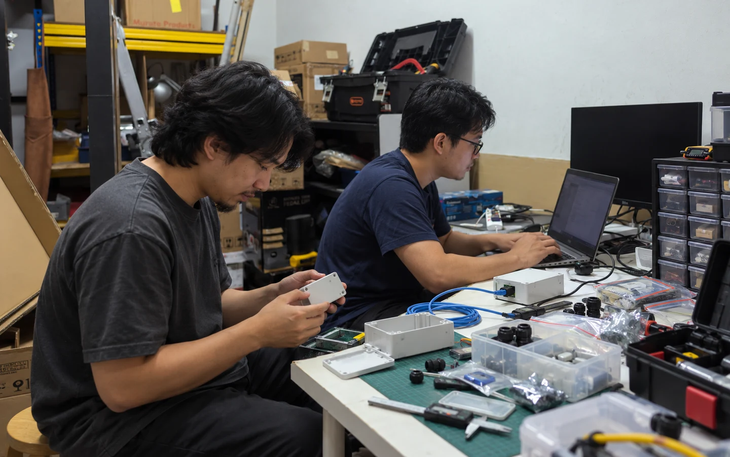 AI-generated illustration grounded in photographs of REKA’s hardware lab. Fictional engineers validate a sensor-to-edge prototype at a working electronics bench; not actual staff, a client deployment or a claimed result.