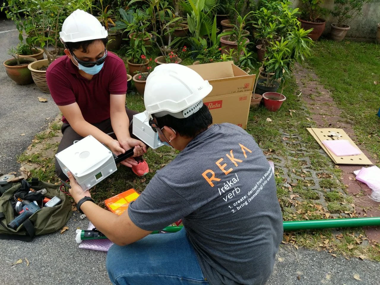 REKA archive photograph. Two people preparing sensing enclosures outdoors in the Murata media kit; no individual identities or specific location assigned.